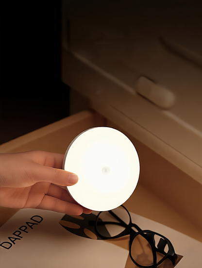 Hand holding a round, glowing light bulb on a desk with a book and glasses in the background.